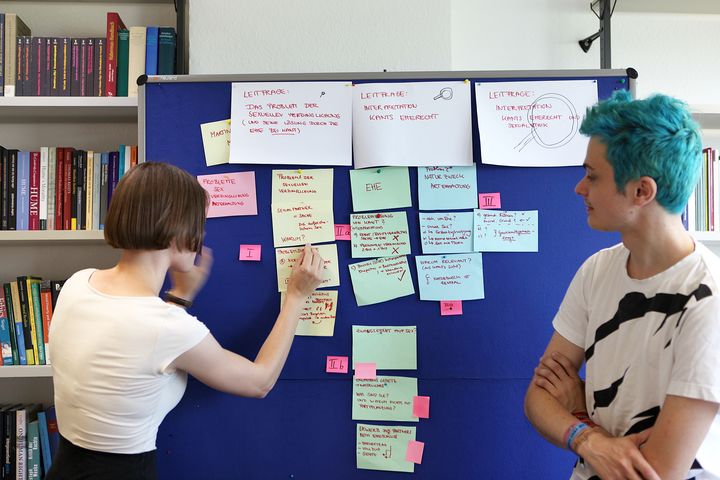 Im Hintergrund eine weiß tapezierte Wand mit einer gebäudestützenden Säule und gefüllten Bücherregalen links und rechts davon. Im Vordergrund eine dunkelblaue Pinnwand. Davor stehen links und rechts 2 Studierende, die beide seitlich bzw. von schräg hinten zu sehen sind. Sie sind mit weißen Shirts bekleidet. Die linke Person befestigt gerade einen gelben Zettel auf der Pinnwand. Insgesamt befinden sich auf dieser 3 große weiße Zettel oben, sowie ein hellroter, 4 gelbe, 6 grüne und 4 hellblaue mittelgroße Zettel spaltenartig unterhalb entsprechend ihrer Farben gruppiert. Ein fünfter gelber Zettel durchbricht dieses Schema und befindet sich, halb durch den ersten weißen Zettel links verdeckt, auf dessen Höhe. Außerdem sind kleine rote, teilweise mit römischen Ziffern beschriftete weitere Zettel über die Pinnwand verteilt, die offenbar eine weitere Gruppierung andeuten sollen. Gut lesbar ist nur die genannte Beschriftung mit römischen Ziffern sowie das Wort "Ehe" auf einem der grünen Zettel.