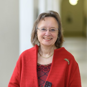 Portrait von Tracy Rosemary. Sie trägt eine rote Bluse und einen roten Mantel und steht lächelnd in einem Gang.